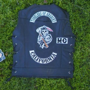 May include: Black leather vest with white patches. The back of the vest features a skull graphic holding a scythe, the words "TODDLERS OF ANARCHY" and "CALIFORNIA". The vest has side laces and the letters "MC".