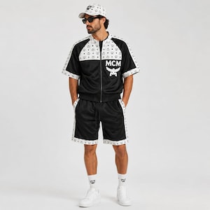 May include: Black and white track suit with a matching baseball cap. The jacket and shorts feature white trim with a repeating logo pattern. The jacket has a zipper closure and the logo "MCM" on the chest.