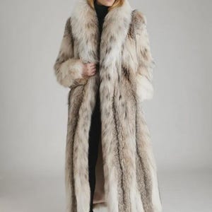 Könnte beinhalten: Ein langer Pelzmantel in Creme-, Braun- und Grautönen. Der Mantel hat einen breiten Kragen und lange Ärmel. Das Model trägt eine schwarze Hose und schwarze Absätze. Der Mantel scheint aus luxuriösem Pelz zu sein.