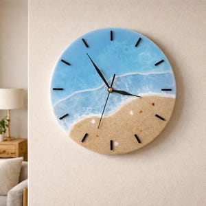 Peut inclure: Une horloge murale ronde avec un motif de scène de plage. La moitié supérieure représente un océan bleu clair avec des vagues blanches, tandis que la moitié inférieure représente une plage de sable. Les aiguilles et les marqueurs d'heure noirs complètent le design. L'horloge mesure environ 30 cm de diamètre.