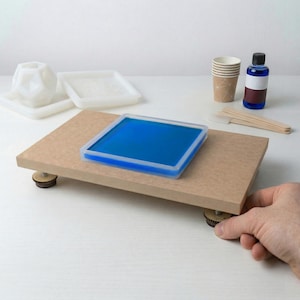 Könnte beinhalten: Ein Bastelprojekt-Setup mit einer Holzplattform, die eine blaue quadratische Harzform hält. Weitere Artikel sind eine weiße geometrische Form, kleine Becher, eine Flasche blaue Flüssigkeit und Rührstäbchen. Die Plattform wird von kleinen, runden Füßen getragen.