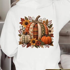 Könnte beinhalten: Ein weißer Kapuzenpullover mit einer großen Grafik auf der Rückseite, die drei Kürbisse mit verschiedenen Mustern wie Karos, Blumen und einfarbigem Orange zeigt. Die Kürbisse sind von Herbstblättern, Sonnenblumen und roten Beeren umgeben.