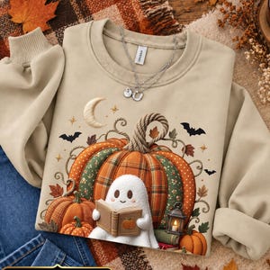 May include: A sand-coloured crewneck sweatshirt featuring a graphic print of a white ghost reading a book in front of a large, patterned pumpkin. The design includes a crescent moon, small stars, bats, and a lantern. A silver necklace rests on the collar.