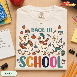 May include: An ivory t-shirt featuring a graphic of three white geese with school supplies and the text BACK TO SCHOOL. The geese are depicted with a backpack, a graduation cap, a scarf, and books. The design includes apples, pencils, and a globe.
