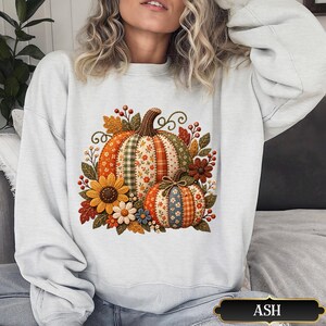 May include: A light grey crewneck sweatshirt featuring a central graphic of two pumpkins with patchwork patterns in orange, green, and blue. The design includes sunflowers, small white flowers, autumn leaves, and red berries surrounding the pumpkins.