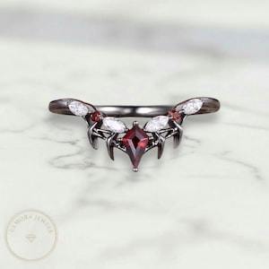 May include: A black metal ring with a central, diamond-shaped, red gemstone. The ring features white and red accent stones and a decorative, curved design. The ring is set against a white marble surface.