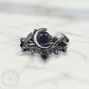 Può includere: Un set di anelli in due pezzi con una fascia argentata scura e un design a mezzaluna. L'anello principale ha una grande pietra blu scuro e il set è adornato con pietre preziose viola più piccole e accenti di foglie. Gli anelli sono su sfondo di marmo bianco.