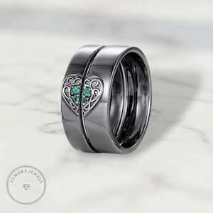 May include: Two matching black rings. One ring features a heart-shaped design with small, green gemstones. The other ring is plain. The rings are displayed on a marble surface. The rings are from Ulmorajewels.