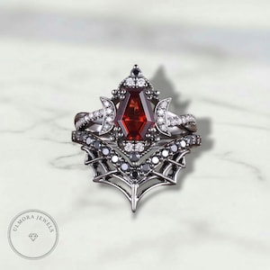 Könnte beinhalten: Ein zweiteiliges Ring-Set mit einem dunklen Metallband und einem großen, facettierten, roten Edelstein. Der obere Ring hat ein Halbmond-Design mit weißen Edelsteinen. Der untere Ring hat ein Spinnennetz-Design mit schwarzen Edelsteinen. Der Markenname "ULMORA JEWELS" befindet sich unten links.