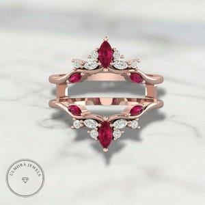 May include: Rose gold ring guard with marquise-cut ruby and diamond accents. The ring features a crown-like design with a central ruby and smaller rubies and diamonds. The ring is designed to be worn with another ring. The brand name "ULMORA JEWELS" is in the bottom left corner.