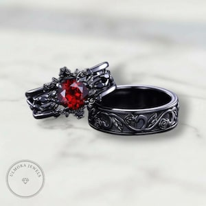 Puede incluir: Dos anillos negros. Uno presenta una gran gema roja y detalles florales intrincados. El otro es una banda simple con un patrón floral. Los anillos están sobre un fondo de mármol blanco.
