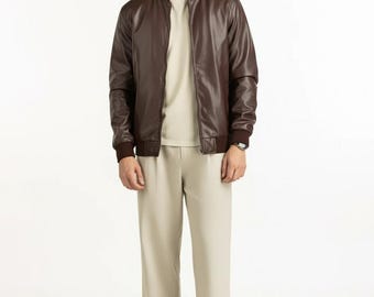 May include: A brown leather bomber jacket with a ribbed collar and cuffs. The jacket is worn over a beige crew neck shirt and paired with beige pants. The outfit is completed with brown dress shoes.
