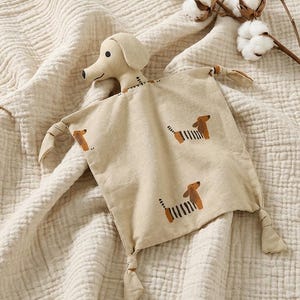 May include: A beige fabric security blanket featuring a plush dachshund head and a square cloth body with knotted corners. The fabric is printed with a repeating pattern of small brown and white dachshunds with striped bodies.