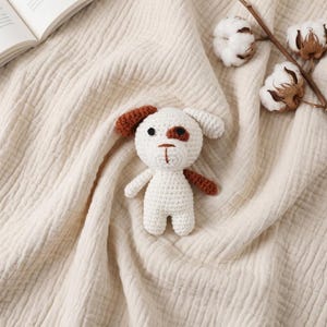 May include: A small, handmade crochet dog toy featuring a white body, a brown patch over one eye, a brown ear, and a white ear. The toy has black embroidered eyes and a brown stitched nose and mouth, crafted with yarn in a simple, textured stitch.