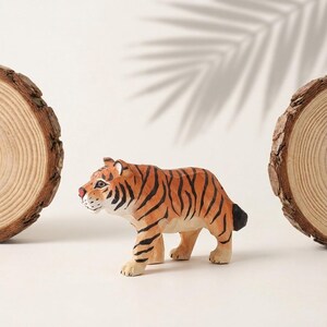 K&ouml;nnte beinhalten: Handgeschnitzte Holz-Tigerfigur mit orangefarbenen und schwarzen Streifen, roter Nase und wei&szlig;en Akzenten. Der Tiger befindet sich in einer Laufhaltung. Zwei Holzscheiben rahmen den Tiger auf einer wei&szlig;en Oberfl&auml;che ein.