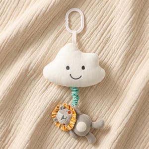 Peut inclure: Un jouet pour bébé en peluche représentant un nuage blanc souriant et un lion gris avec une crinière orange et jaune. Le jouet est attaché à un anneau en plastique blanc avec un tissu vert enroulé. Le fond est un tissu texturé de couleur crème.