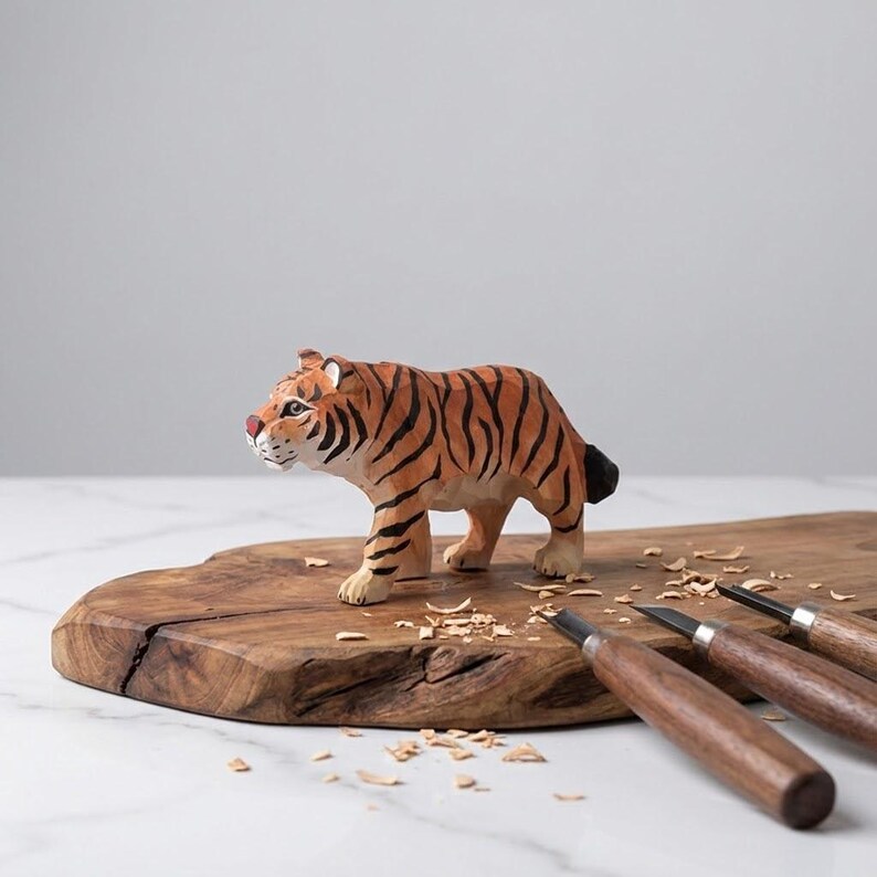 K&ouml;nnte beinhalten: Handgeschnitzte Holz-Tigerfigur mit orangefarbenen und schwarzen Streifen, die auf einem Holzschneidebrett steht. Der Tiger hat ein wei&szlig;es Gesicht und wei&szlig;e Pfoten. Schnitzwerkzeuge mit Holzgriffen sind ebenfalls zu sehen.