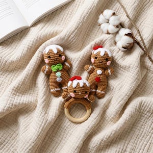 Könnte beinhalten: Zwei braune gehäkelte Lebkuchenpuppen und eine Lebkuchenrassel mit Holzring. Die Puppen haben weiße Glasurdetails, rosa Wangen und kleine bunte Knöpfe. Die Rassel hat ein rotes Mützendetail, während die Puppen grüne oder rote Akzente aufweisen.