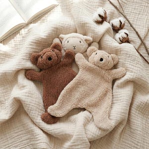 May include: Three plush security blankets featuring animal heads: a dark brown bear, a light beige bear, and a cream-colored lamb. Each toy has a soft, textured fleece-like fabric body with outstretched arms and legs, arranged on a cream muslin cloth.