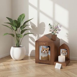 Puede incluir: Una casa para gatos de madera con forma de casa, con una foto enmarcada de un gato jengibre. A su lado hay un marco de madera arqueado más pequeño con una vela y otra foto de gato. Una planta en maceta y flores también están en la imagen.