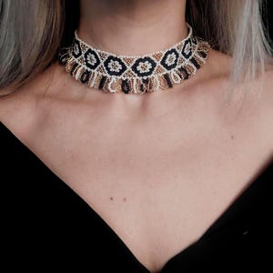 May include: A beaded choker necklace with a geometric pattern. The necklace features a repeating design of black, gold, and white beads. The choker is worn around the neck, and the background is black.