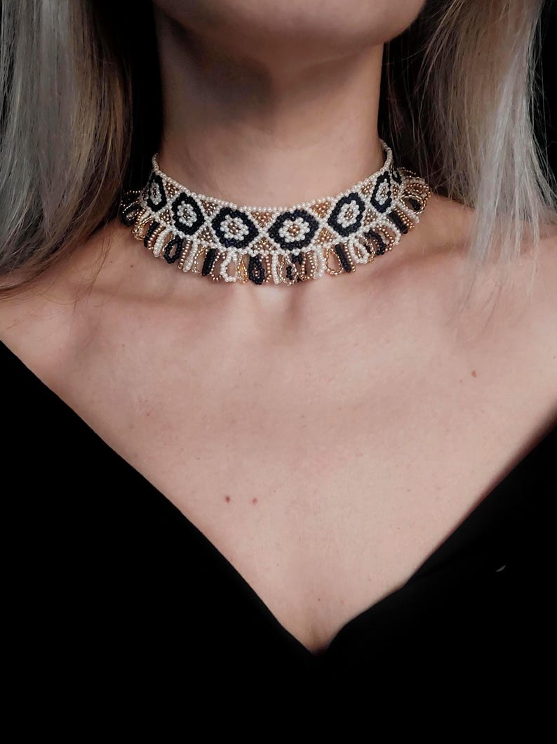 May include: A beaded choker necklace with a geometric pattern. The necklace features a repeating design of black, gold, and white beads. The choker is worn around the neck, and the background is black.