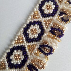 May include: Close-up of a beaded bracelet featuring a geometric pattern. The bracelet is made with small, round beads in white, gold, and deep purple. The design includes circular and floral motifs, creating a textured and ornate appearance.