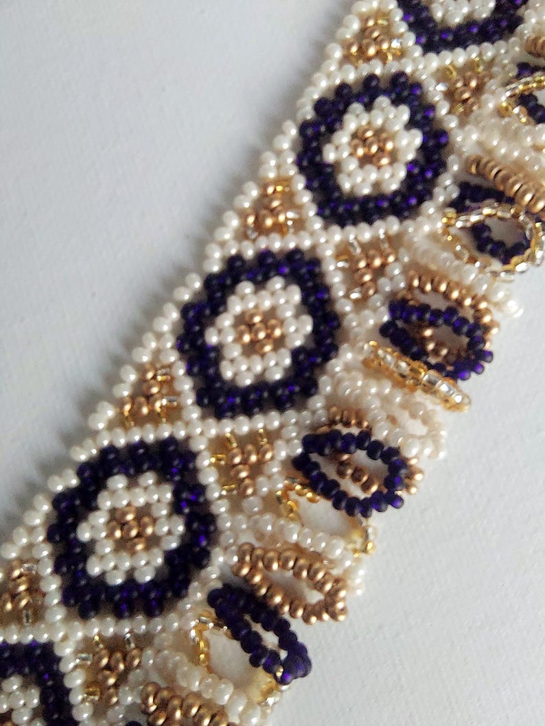 May include: Close-up of a beaded bracelet featuring a geometric pattern. The bracelet is made with small, round beads in white, gold, and deep purple. The design includes circular and floral motifs, creating a textured and ornate appearance.