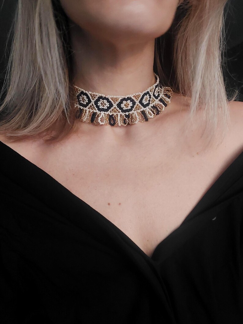 May include: A beaded choker necklace with a geometric pattern. The necklace features a repeating design of black, gold, and white beads. The choker is worn around the neck and is set against a black background.