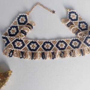May include: A beaded choker necklace featuring a geometric pattern of navy blue and gold beads. The necklace has a fringe of gold beads along the bottom edge and a gold chain with a star charm.
