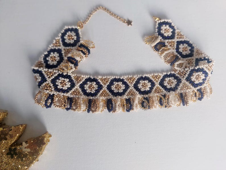 May include: A beaded choker necklace featuring a geometric pattern of navy blue and gold beads. The necklace has a fringe of gold beads along the bottom edge and a gold chain with a star charm.