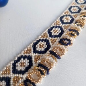 May include: Close-up of a beaded bracelet with a geometric pattern. The bracelet features navy blue, gold, and white beads arranged in a repeating design. The beads are small and create a textured surface. The bracelet is likely handmade.