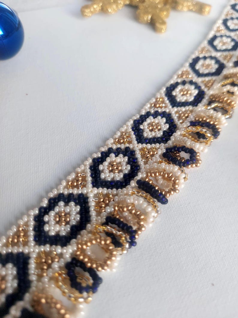 May include: Close-up of a beaded bracelet with a geometric pattern. The bracelet features navy blue, gold, and white beads arranged in a repeating design. The beads are small and create a textured surface. The bracelet is likely handmade.