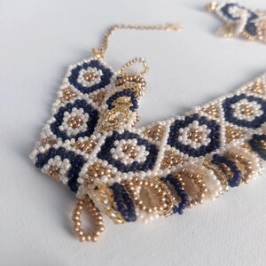 May include: A beaded bracelet with a geometric pattern. The bracelet features navy blue, white, and gold beads. The design includes floral and diamond shapes. The bracelet is on a white surface.