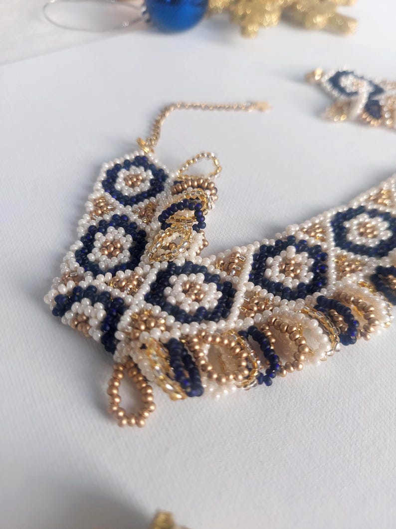 May include: A beaded bracelet with a geometric pattern. The bracelet features navy blue, white, and gold beads. The design includes floral and diamond shapes. The bracelet is on a white surface.