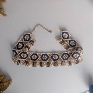 May include: A beaded choker necklace with a geometric pattern in navy blue, gold, and cream. The necklace has a fringe detail along the bottom edge and a delicate chain closure. The design features repeating floral-like shapes.
