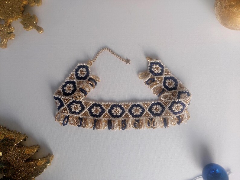 May include: A beaded choker necklace with a geometric pattern in navy blue, gold, and cream. The necklace has a fringe detail along the bottom edge and a delicate chain closure. The design features repeating floral-like shapes.
