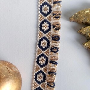 May include: Close-up of a beaded trim with a repeating pattern of navy blue and gold geometric shapes on a cream background. The trim is likely used for embellishing clothing or accessories. The beads are small and tightly woven.