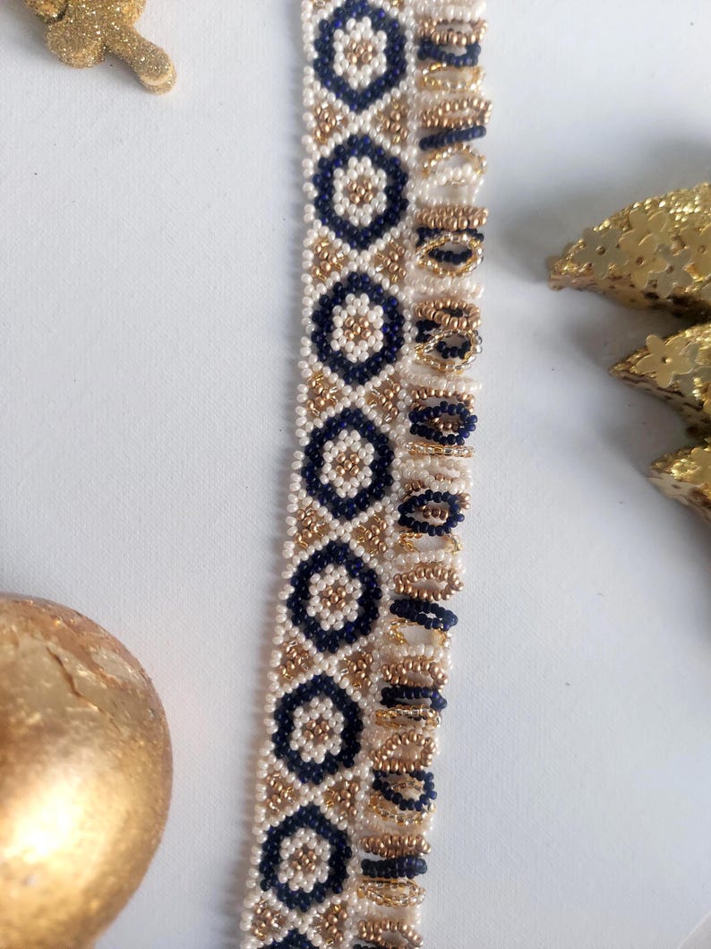 May include: Close-up of a beaded trim with a repeating pattern of navy blue and gold geometric shapes on a cream background. The trim is likely used for embellishing clothing or accessories. The beads are small and tightly woven.