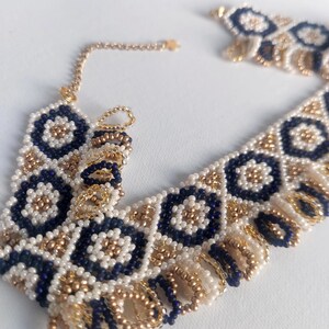 May include: A beaded necklace featuring a geometric pattern of navy blue, white, and gold beads. The necklace has a delicate gold chain and a fringe detail along the bottom edge. The design is symmetrical and intricate, with a bohemian aesthetic.
