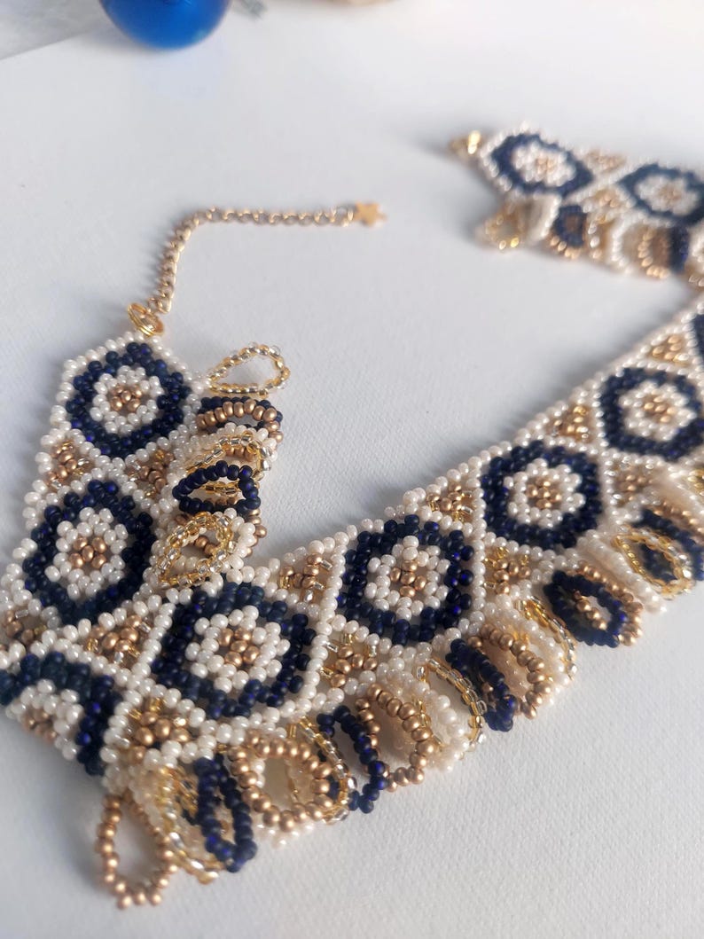 May include: A beaded necklace featuring a geometric pattern of navy blue, white, and gold beads. The necklace has a delicate gold chain and a fringe detail along the bottom edge. The design is symmetrical and intricate, with a bohemian aesthetic.
