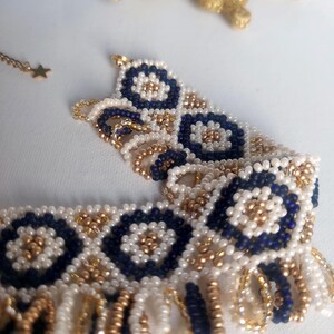 May include: A beaded bracelet with a floral design in navy blue, white, and gold. The bracelet is made of small beads and has a fringe detail. A gold star-shaped charm hangs from a delicate chain.