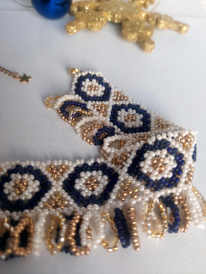 May include: A beaded bracelet with a floral design in navy blue, white, and gold. The bracelet is made of small beads and has a fringe detail. A gold star-shaped charm hangs from a delicate chain.