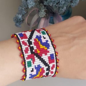 Könnte beinhalten: Ein breites, perlenbesetztes Armband mit einem farbenfrohen geometrischen Muster. Das Armband hat rote, weiße, schwarze, blaue und gelbe Perlen. Die Ränder sind mit kleinen roten und gelben Perlen verziert. Das Armband ist etwa 5 cm breit.
