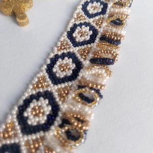 May include: Close-up of a beaded bracelet with a repeating pattern of navy blue and gold floral designs on a white background. The bracelet is made of small beads. A gold glittery ornament is in the background.