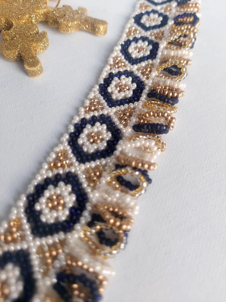 May include: Close-up of a beaded bracelet with a repeating pattern of navy blue and gold floral designs on a white background. The bracelet is made of small beads. A gold glittery ornament is in the background.