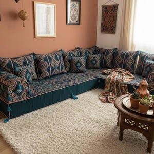 Op de afbeelding: Een grote hoekbank met turquoise en patroon kussens en basis. De bank staat in een woonkamer met een wit tapijt, een houten salontafel en decoratieve wandkunst. Een patroon deken ligt op de bank.