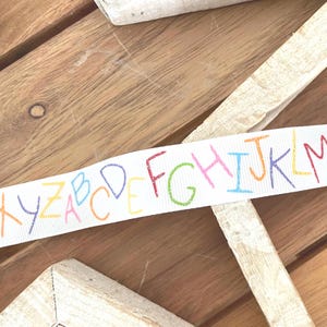 May include: White ribbon featuring the alphabet in a rainbow of colours. The letters are hand-drawn in red, yellow, green, and blue. The ribbon is laid on a wooden surface.