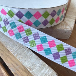 May include: White ribbon with a repeating pattern of colorful, glittery diamonds. The diamonds are in shades of pink, green, purple, and teal. The ribbon is on a wooden surface.