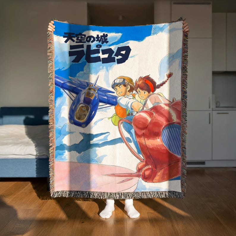 A person is holding up a colorful woven blanket with an illustration of two characters on a plane against a cloudy sky background.
